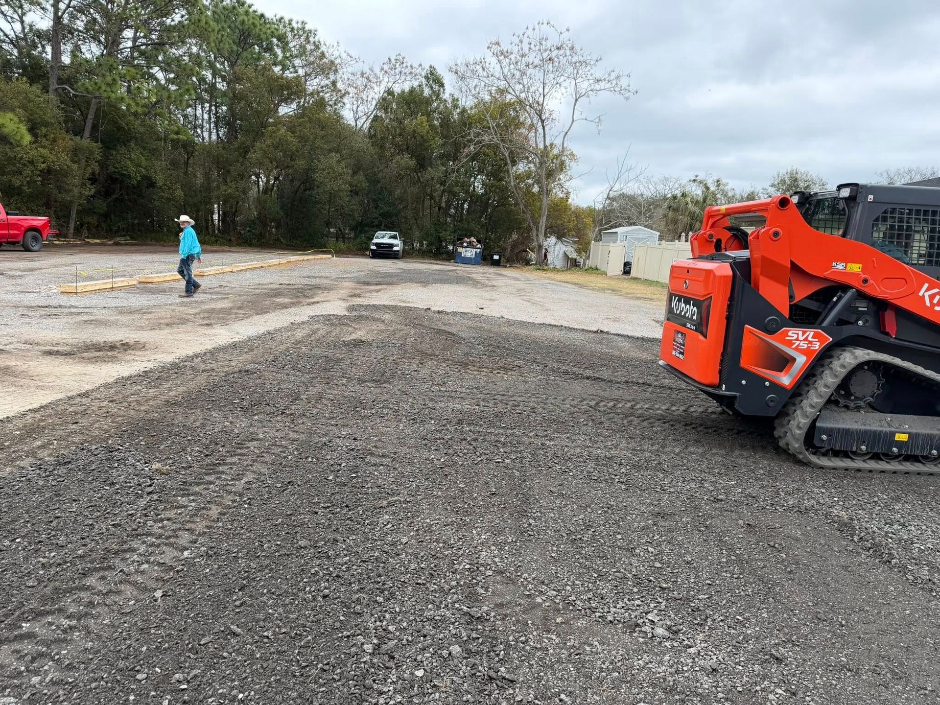 An orange skid steer on a gravel lot near a person walking and parked vehicles under a cloudy sky.