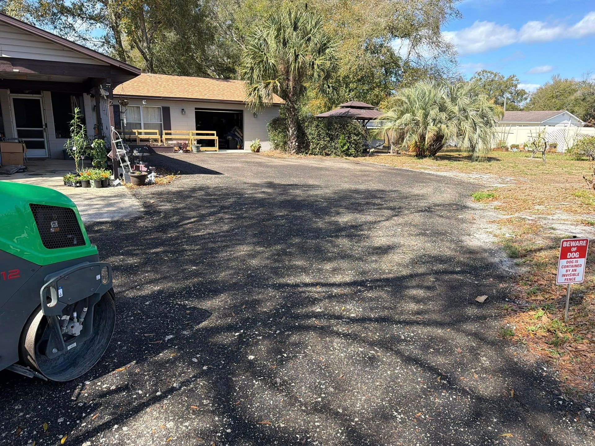 A green steamroller on a newly paved, dark asphalt driveway leading toward a house and detached garage on a sunny day.