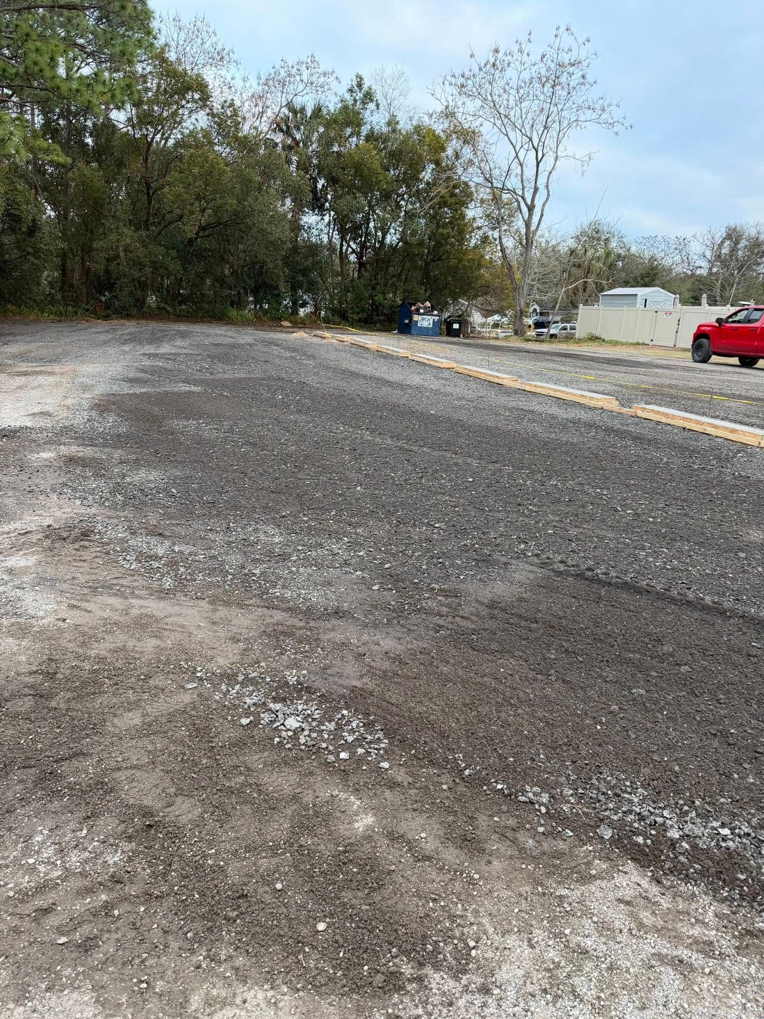 A freshly paved black asphalt area transitions into a light gravel surface, bordering trees and a parked red truck.