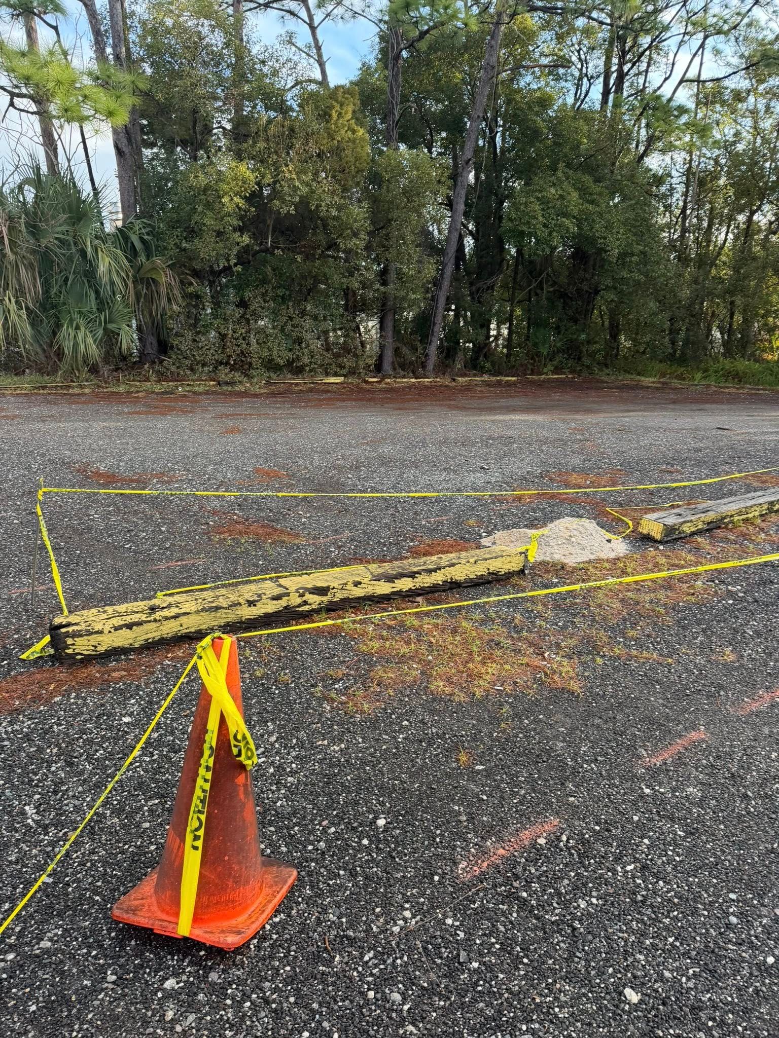 An orange traffic cone and yellow caution tape demarcate a construction or repair area on an asphalt parking lot.