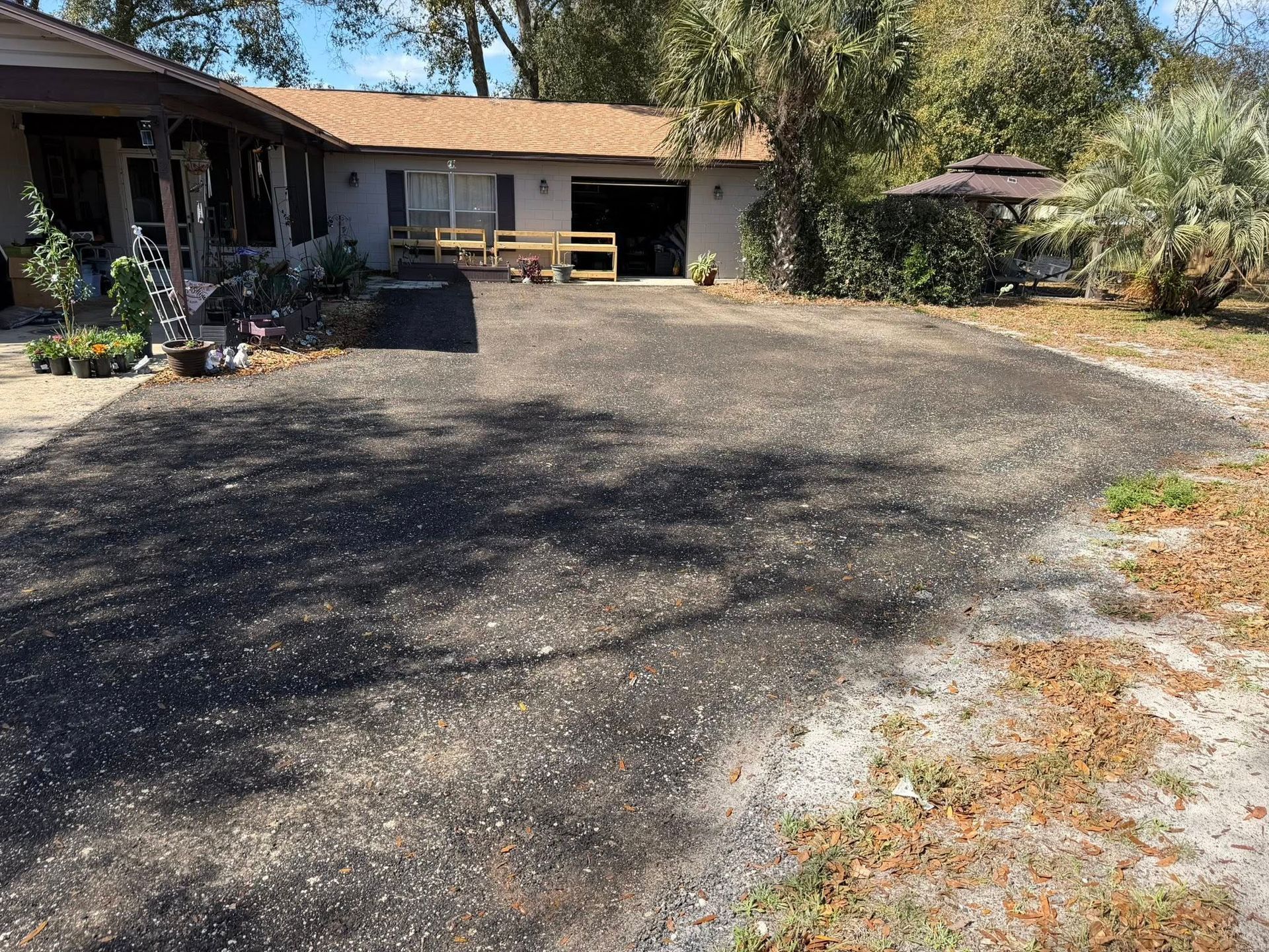 A wide, dark asphalt driveway leads to a single-story home with a garage, bordered by trees and landscaping.