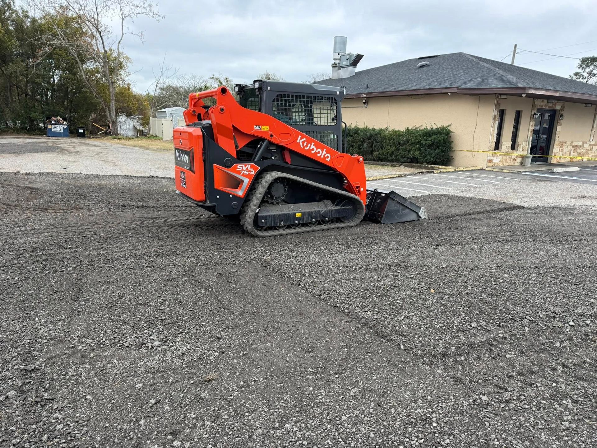 An orange Kubota compact track loader sits on a gravel lot in front of a tan building.