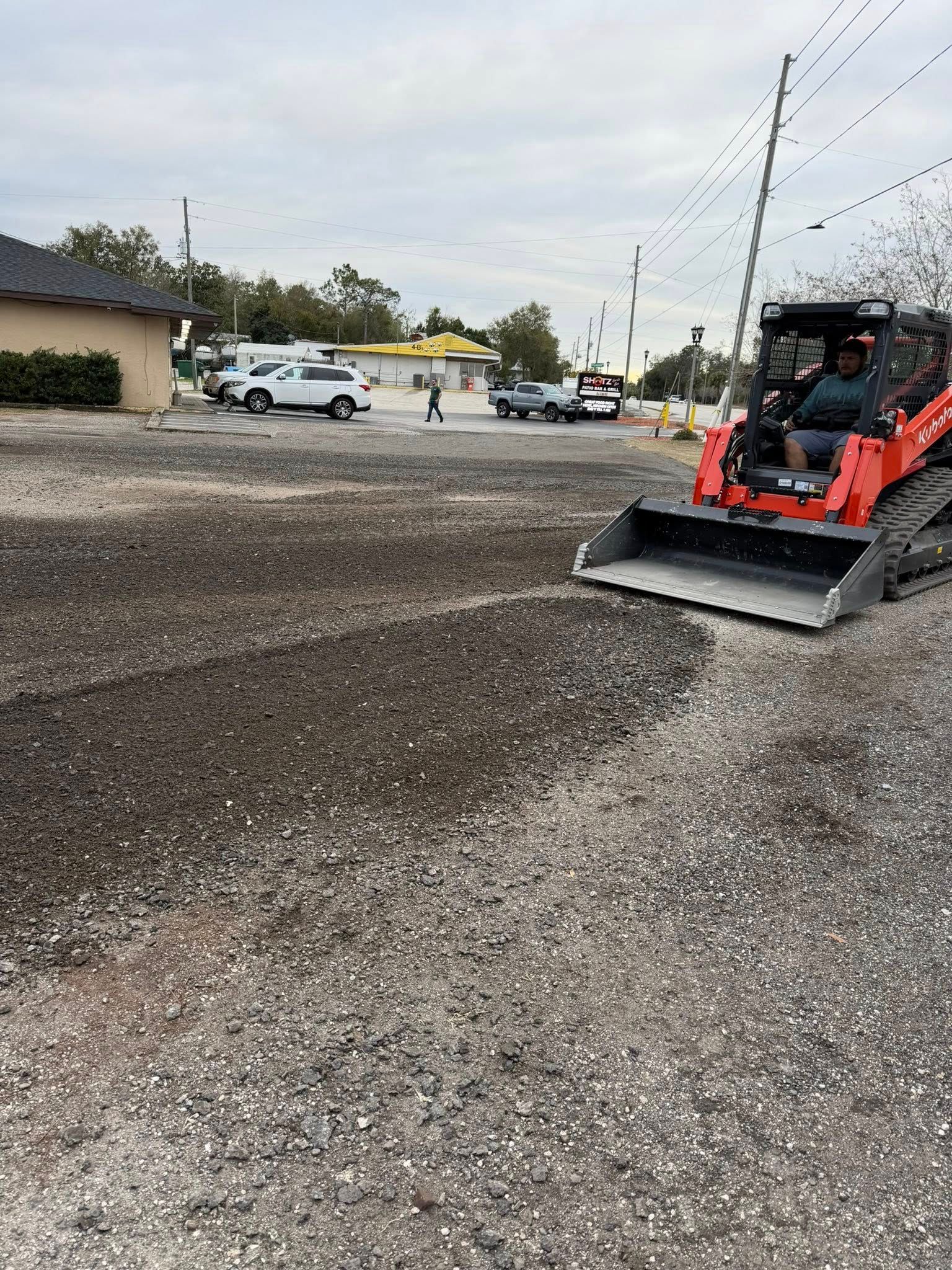 A bright orange skid steer loader grading a gravel lot on an overcast day with buildings and vehicles in the background.