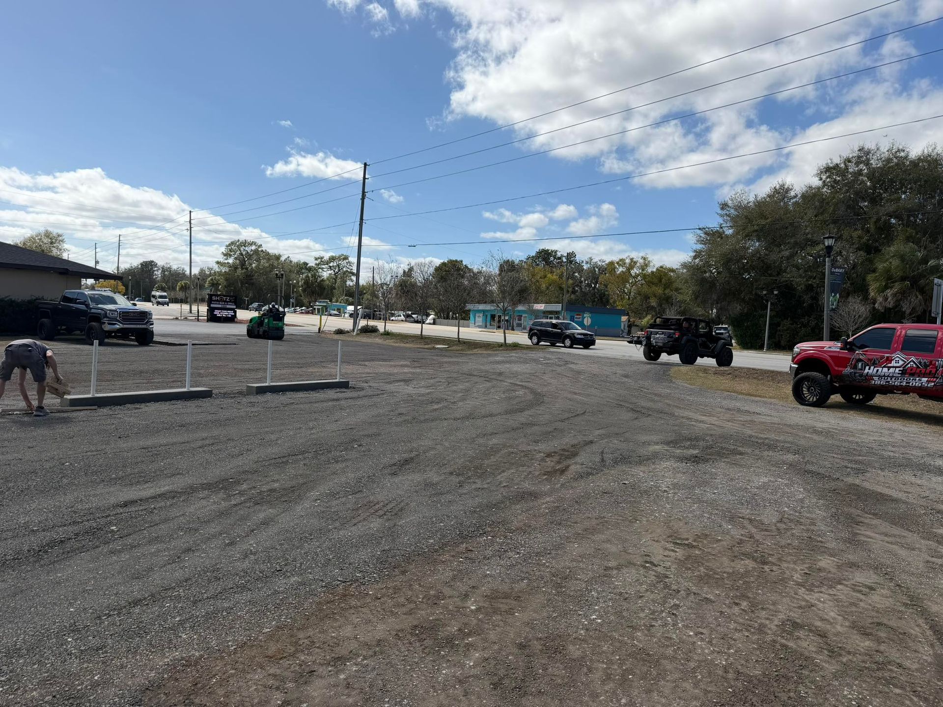 A gravel parking lot under a blue sky, featuring a person working on the left and several parked vehicles in the distance.