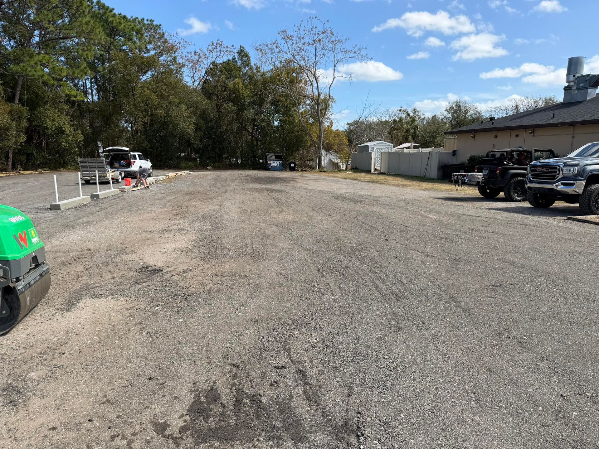 A gravel parking lot under a blue sky, featuring a green roller in the foreground and parked vehicles in the background.