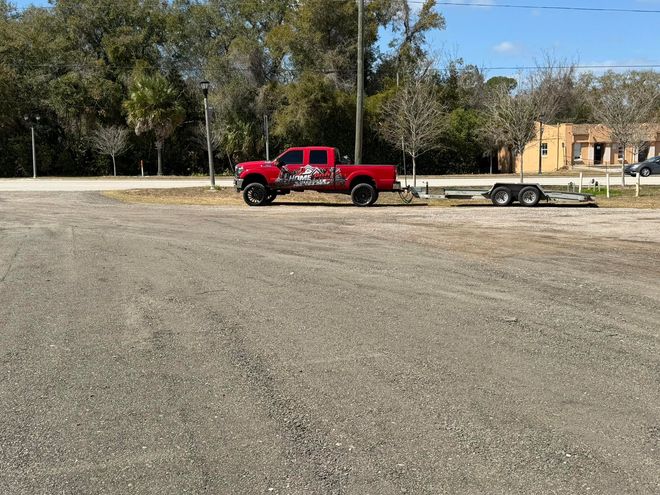 A red pickup truck pulling an empty flatbed trailer parked on a gravel lot next to trees and a small building.