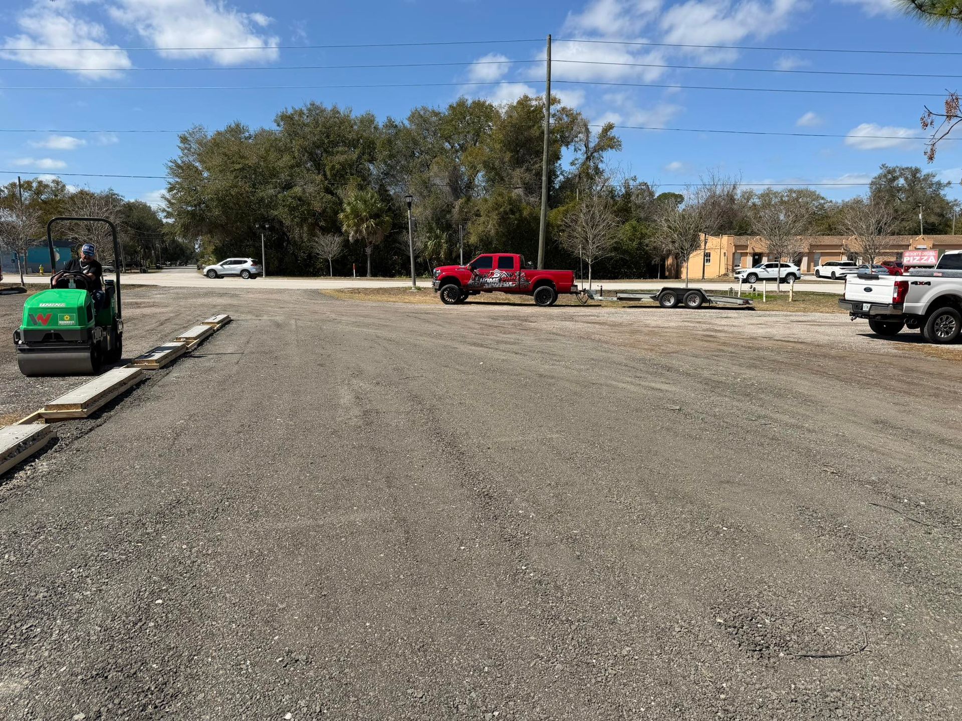 A construction worker on a small green roller flattens a gravel lot with parked trucks nearby under a blue sky.