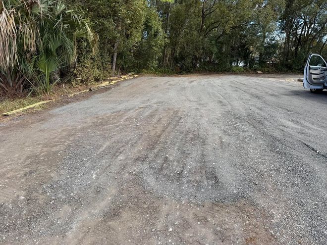 An unpaved gravel lot bordered by trees and dense greenery, with the back of a vehicle visible on the right.