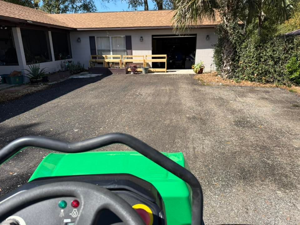 A view from the driver's seat of a green ride-on mower facing a house with a gravel driveway and a wooden barrier.