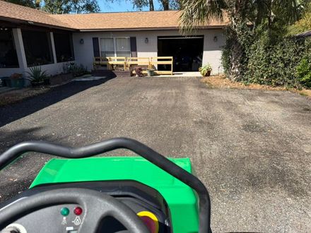 A view from the driver's seat of a green ride-on mower facing a house with a gravel driveway and a wooden barrier.