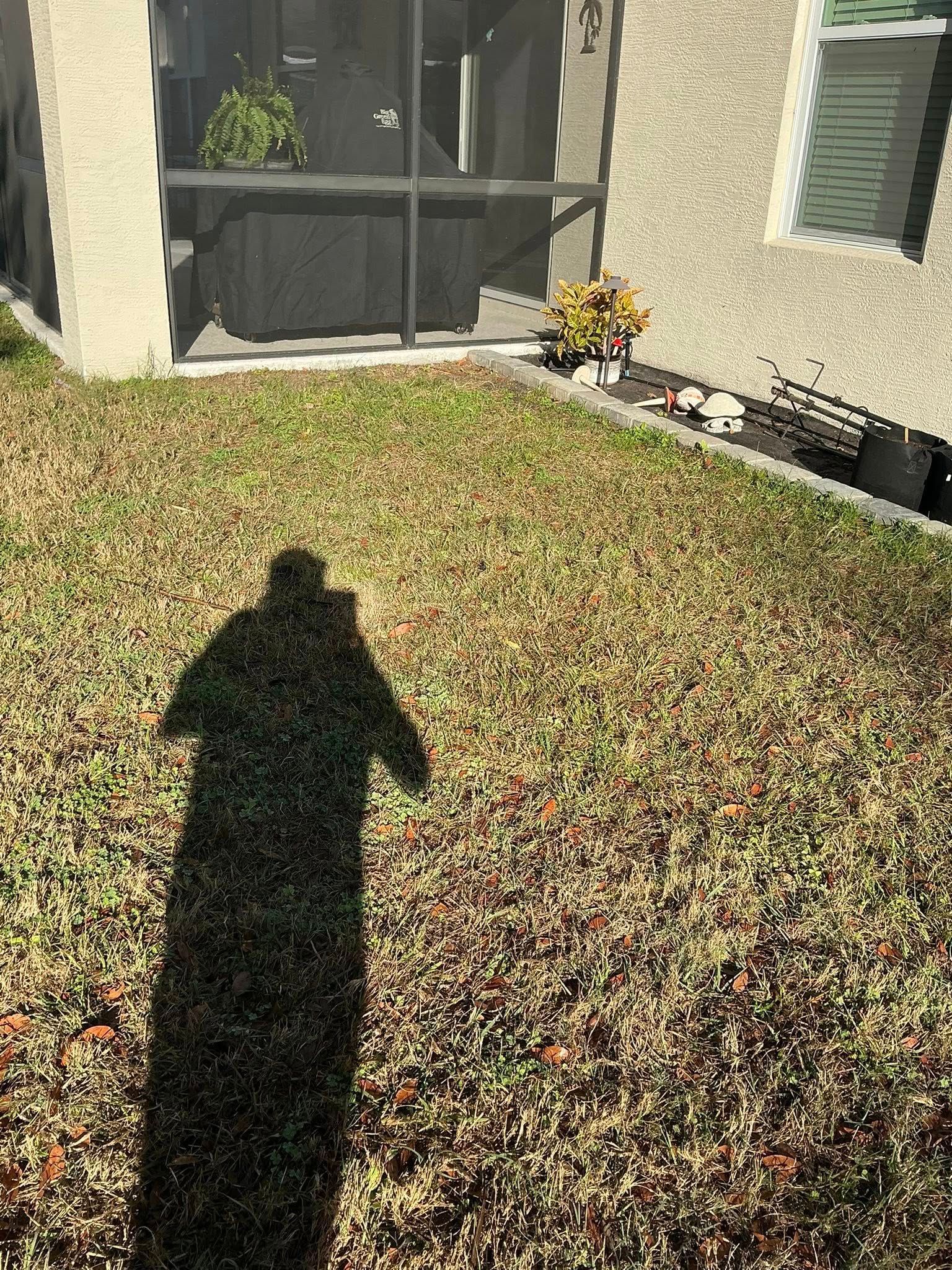 A person’s long shadow stretches across a dry, grassy backyard toward a screened patio and the side of a house.