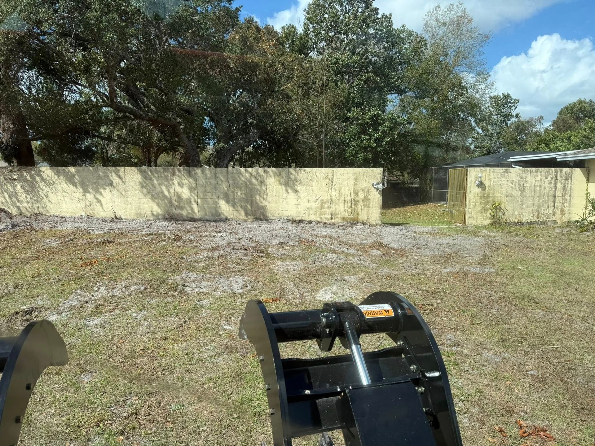 View from a vehicle over a grassy dirt lot toward a pale wall and trees under a blue sky