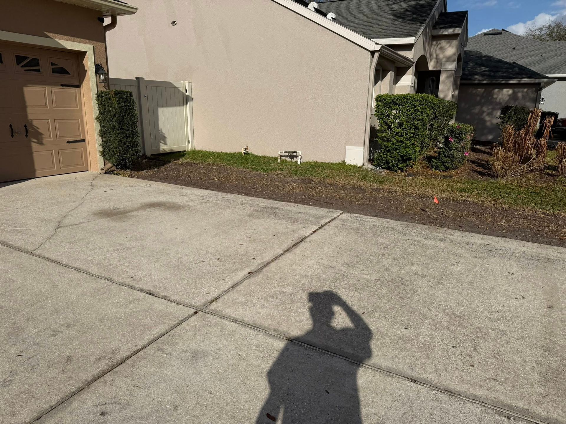 A concrete driveway leads to a garage next to a beige house with a small yard and a shadow of a person in the foreground.