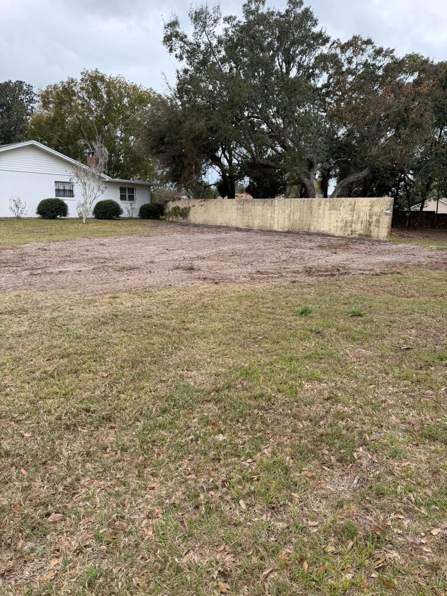 A grass yard with a large patch of gravel in front of a white house and a low stone wall under cloudy skies.
