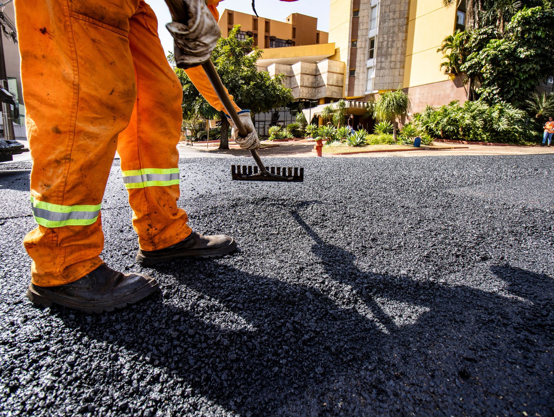 Worker spreading asphalt on a driveway with a rake, wearing orange safety pants and boots