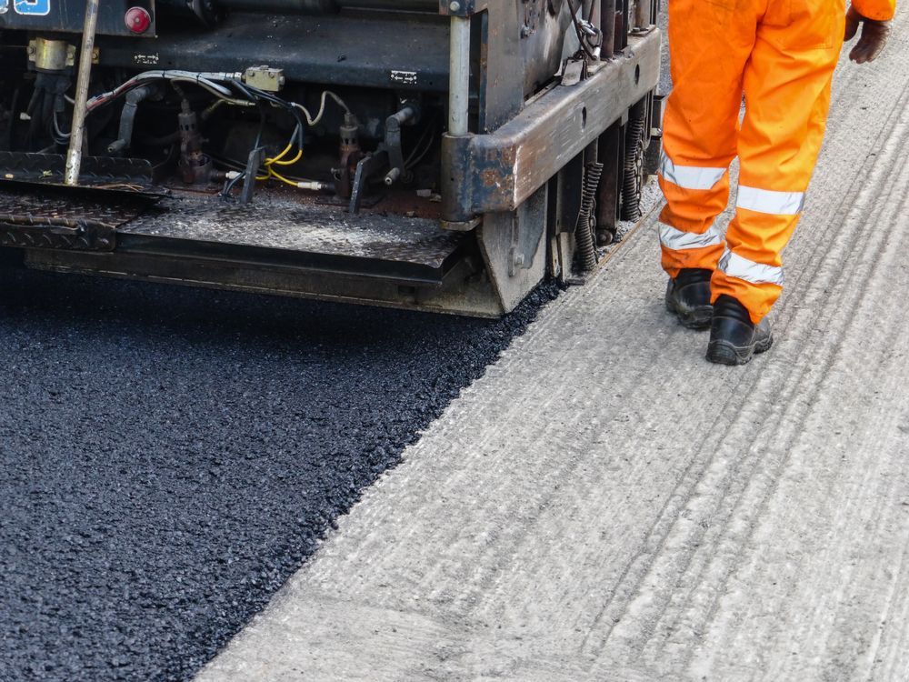 Road paving machine laying fresh asphalt beside a worker in orange safety gear