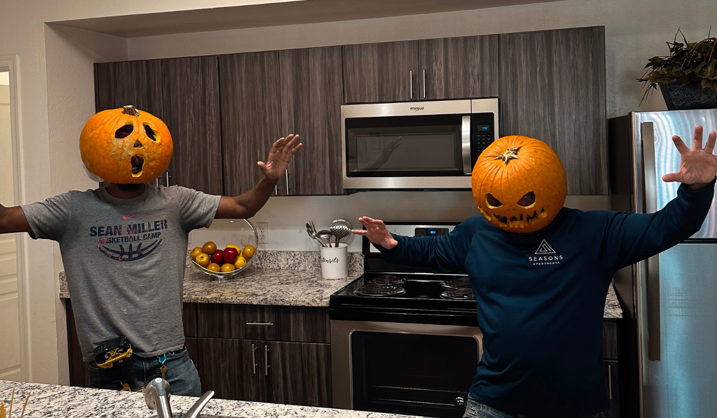 Two male residents in the kitchen wearing carved pumpkin heads.