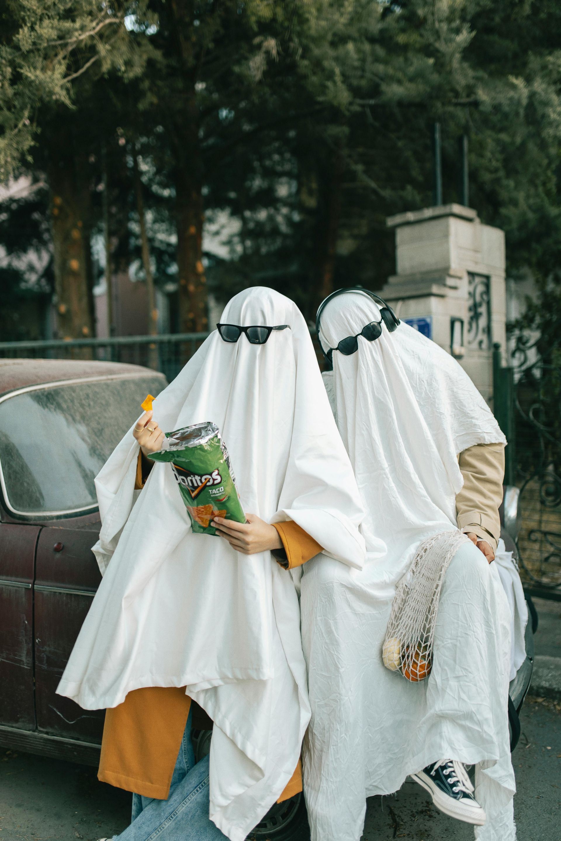 Have some Halloween fun with your roommates! Two people in sheet ghost costumes wear sunglasses and snack on chips.