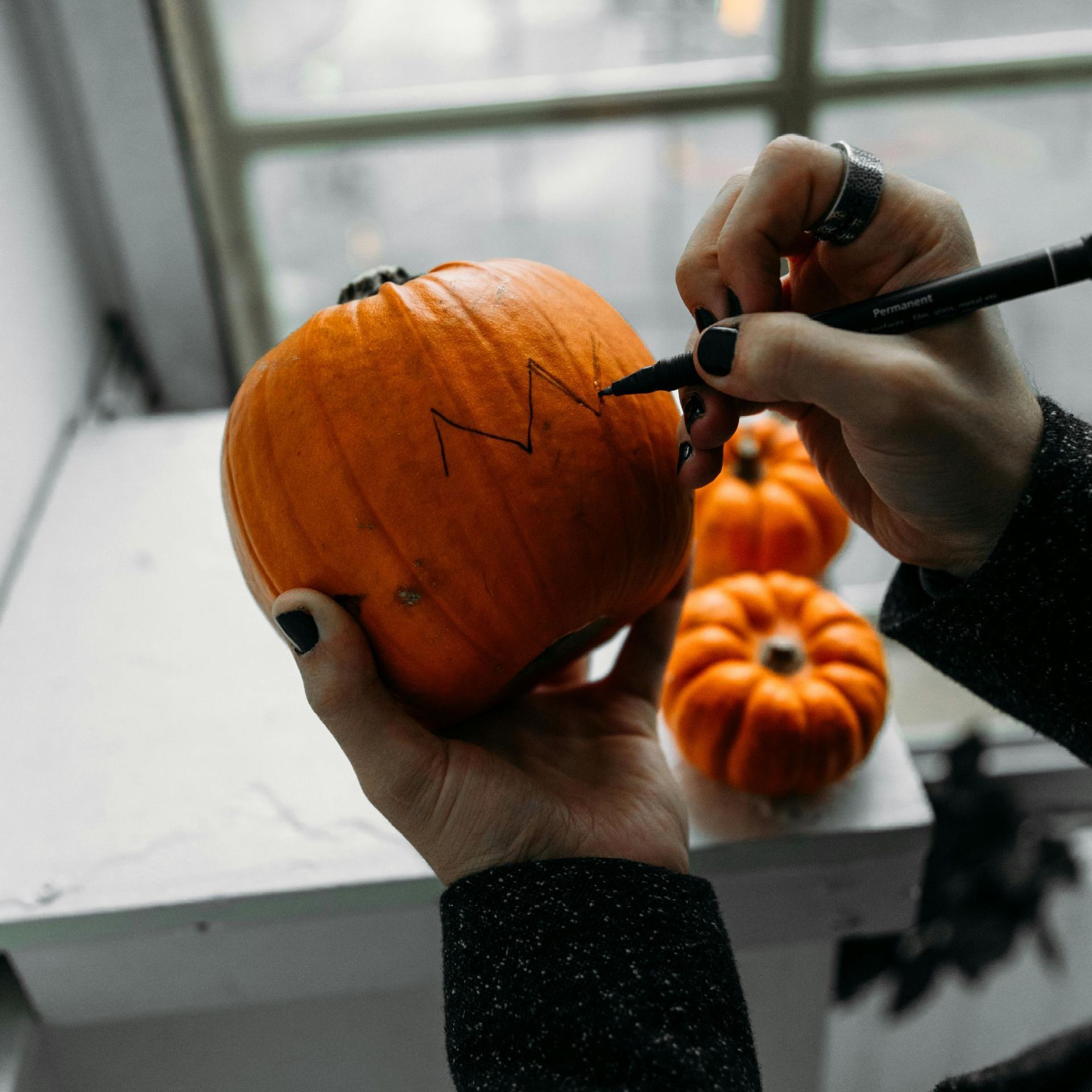 Two hands can be seen drawing a design on a pumpkin with two other pumpkins in the background.
