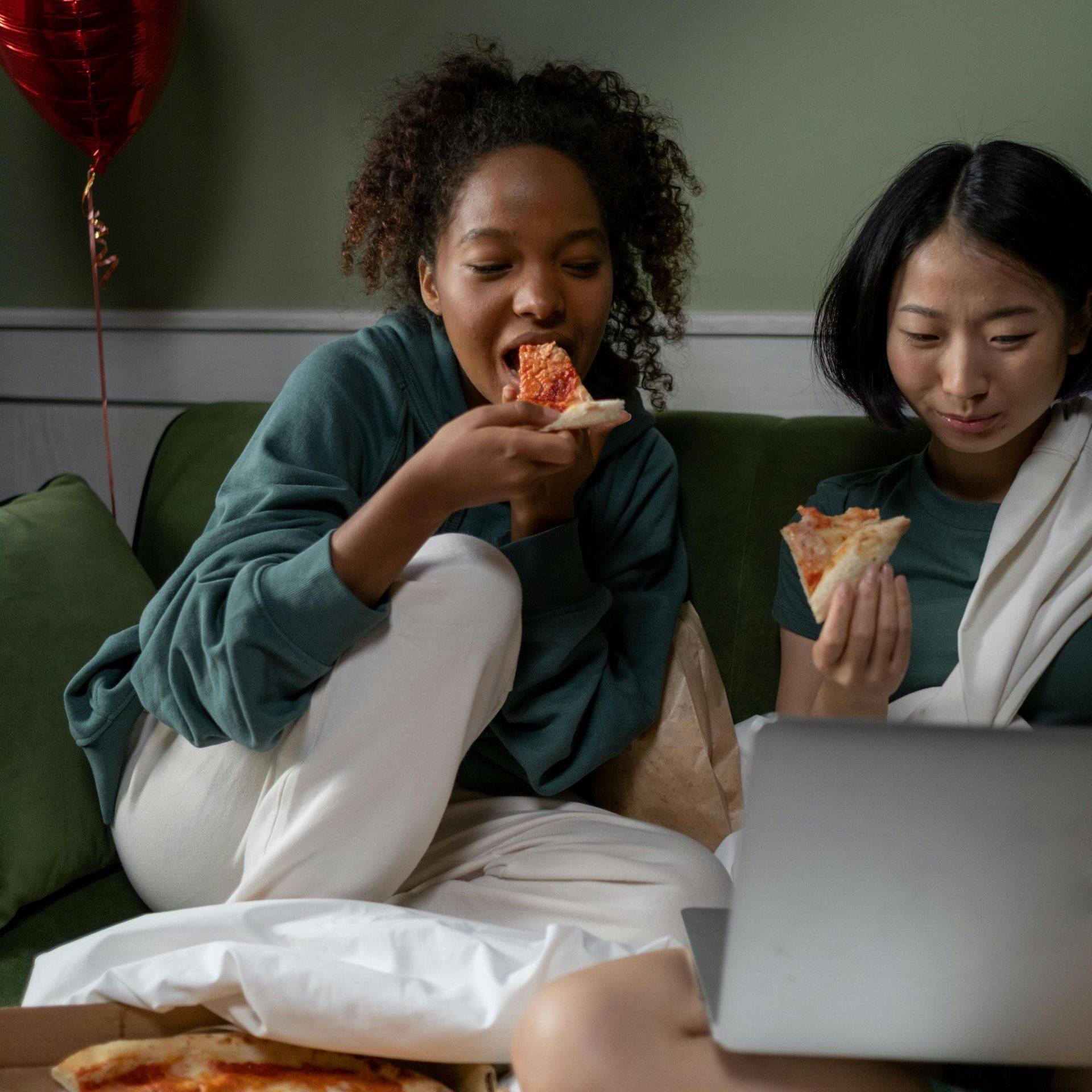 Grab some popcorn and hot cocoa at the Clubhouse! Two females are seen eating pizza and watching a laptop. They are sitting on a green couch with blankets.