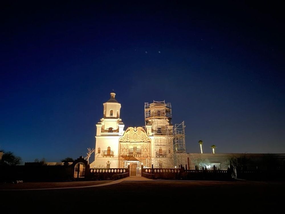 The historic San Xavier church illuminated, with silhouettes of visitors and scaffolding on the tower.