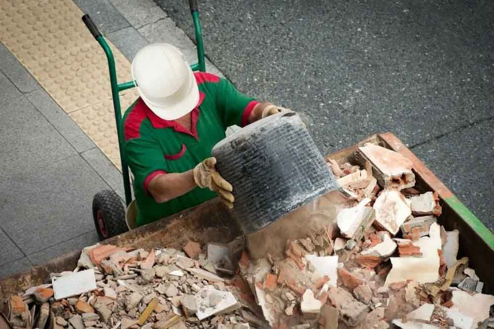 Worker dumping concrete and brick debris into a metal dumpster.