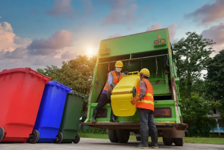 Two workers loading a yellow bin into a green garbage truck.
