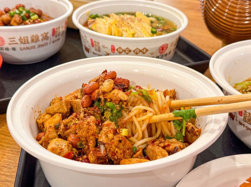 Bowl of spicy noodle salad with chopsticks, surrounded by other takeaway bowls on a wooden table