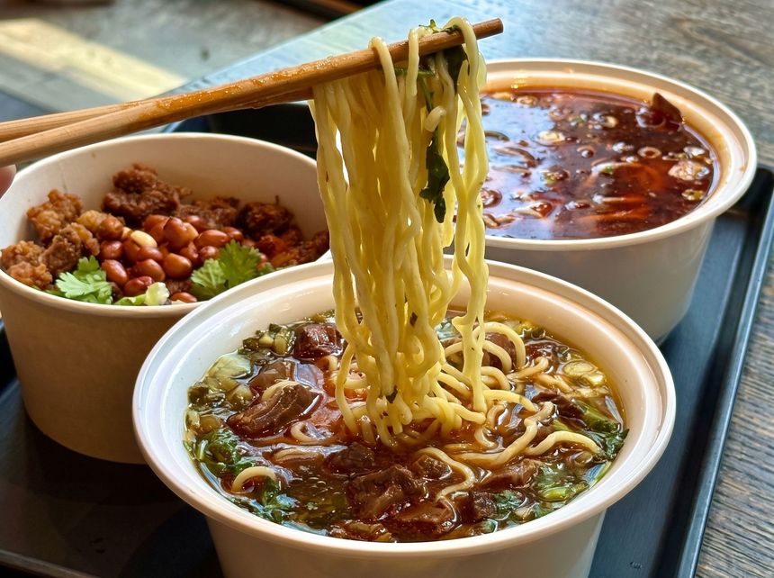 Three bowls of steaming noodle soup, one lifted with chopsticks, on a table outdoors.