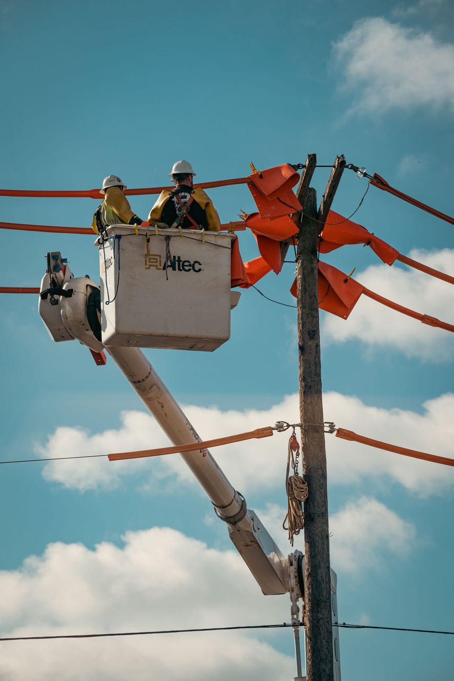 Two workers in a bucket lift adjusting orange streamers on a pole against a blue sky.