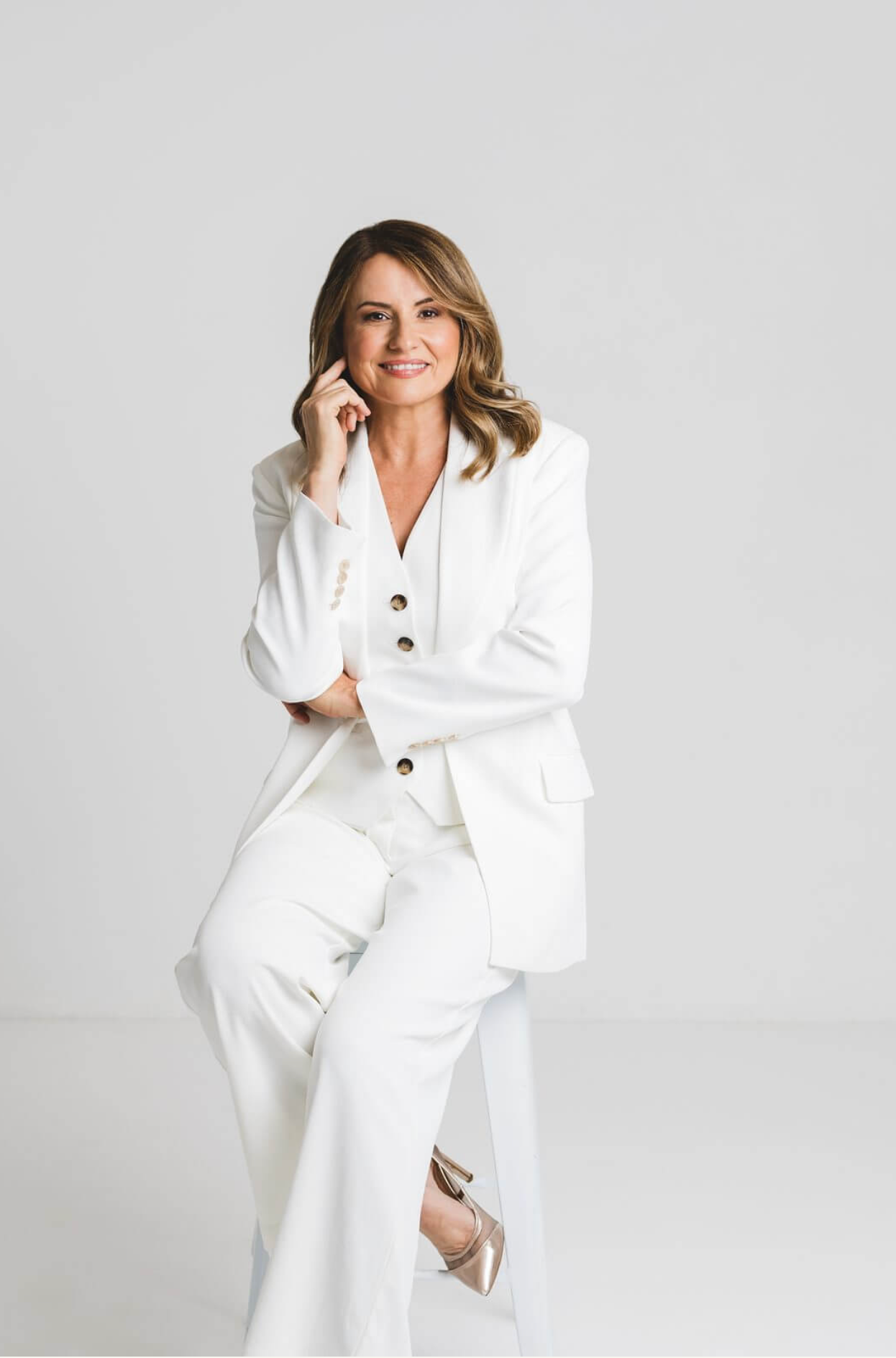 Woman in a white suit seated on a stool against a light gray studio backdrop.