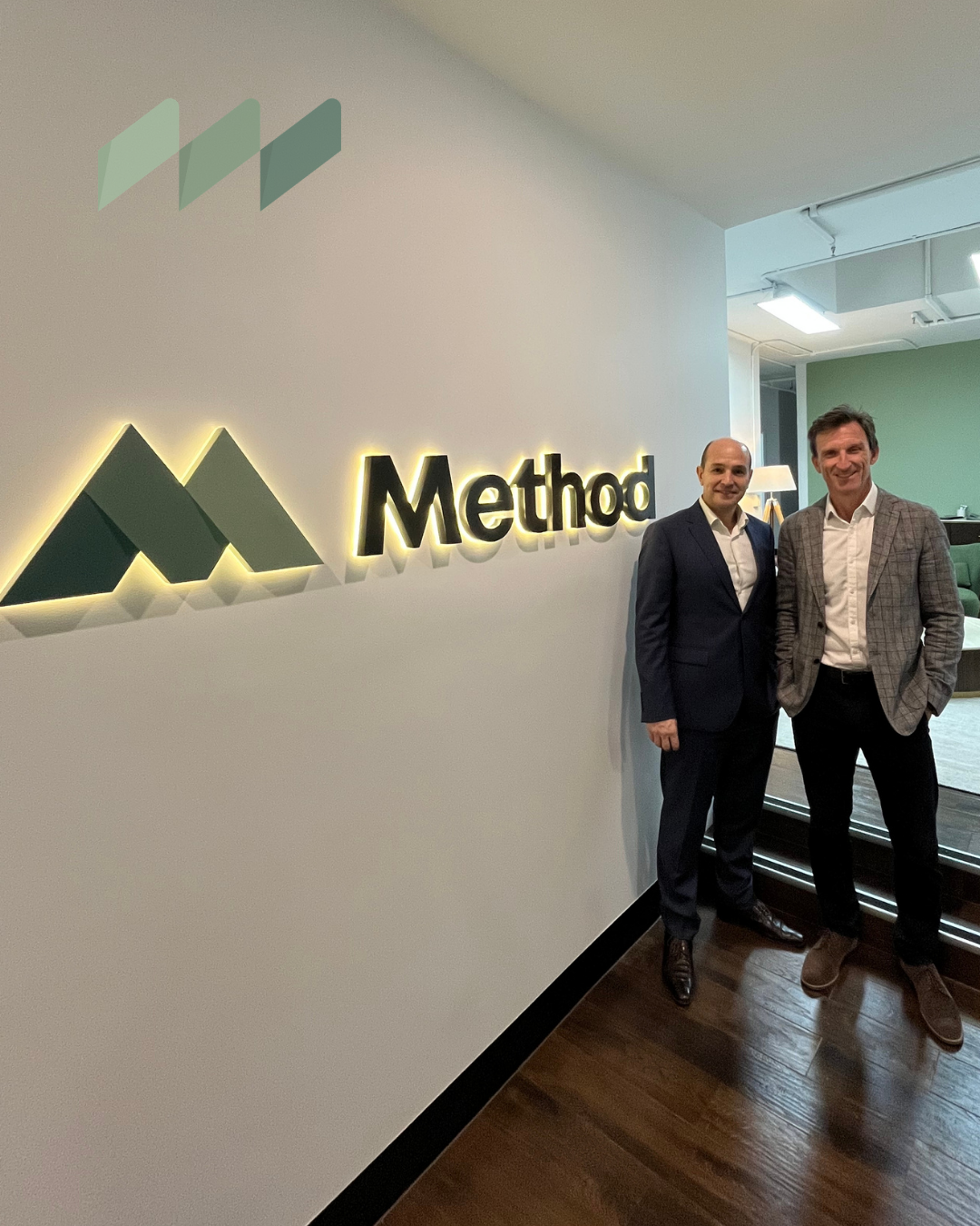 Two men standing in a modern office lobby beside a lit Method logo on the wall