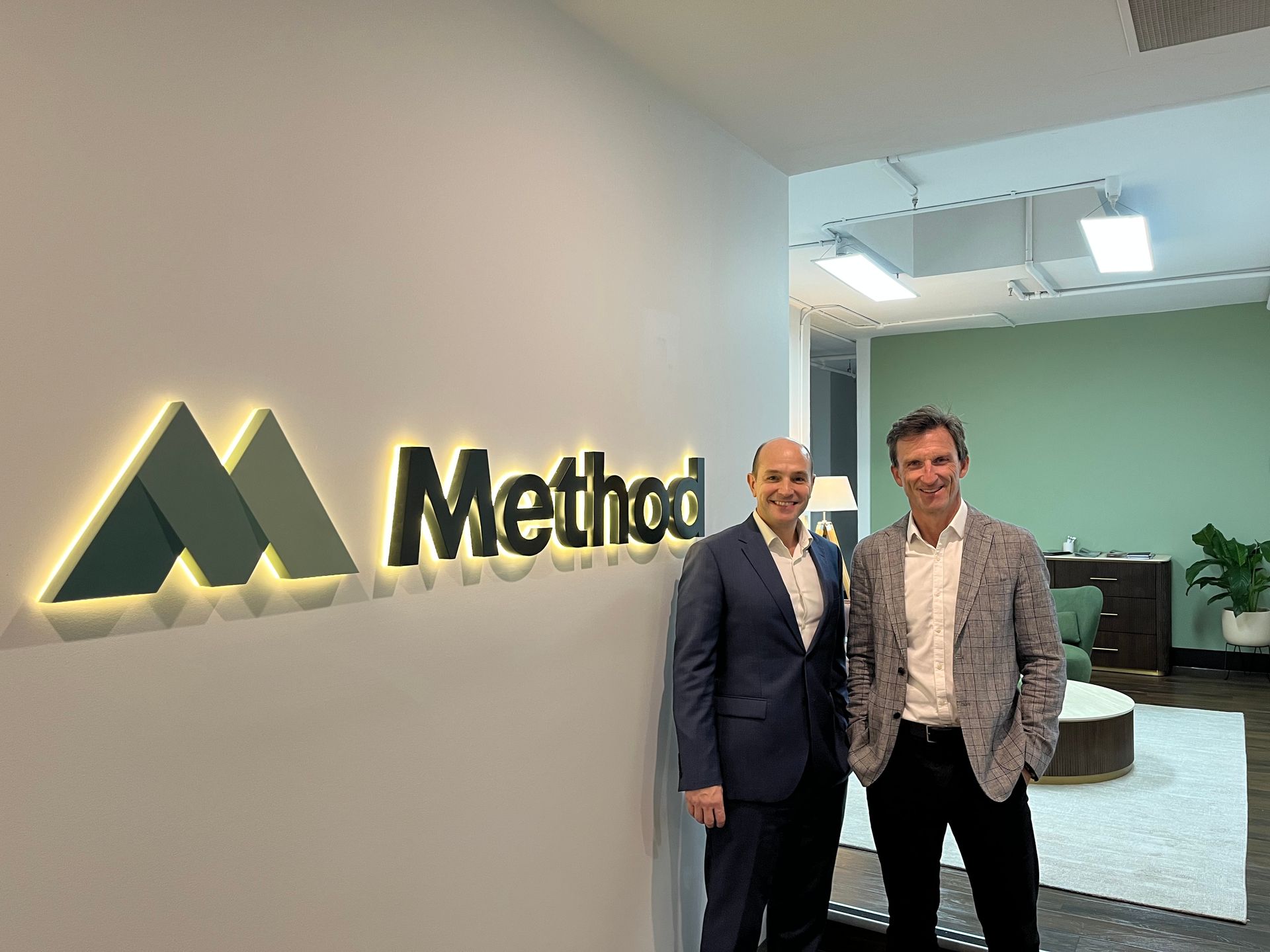 Two men standing in a lobby beside a lit Method logo wall sign