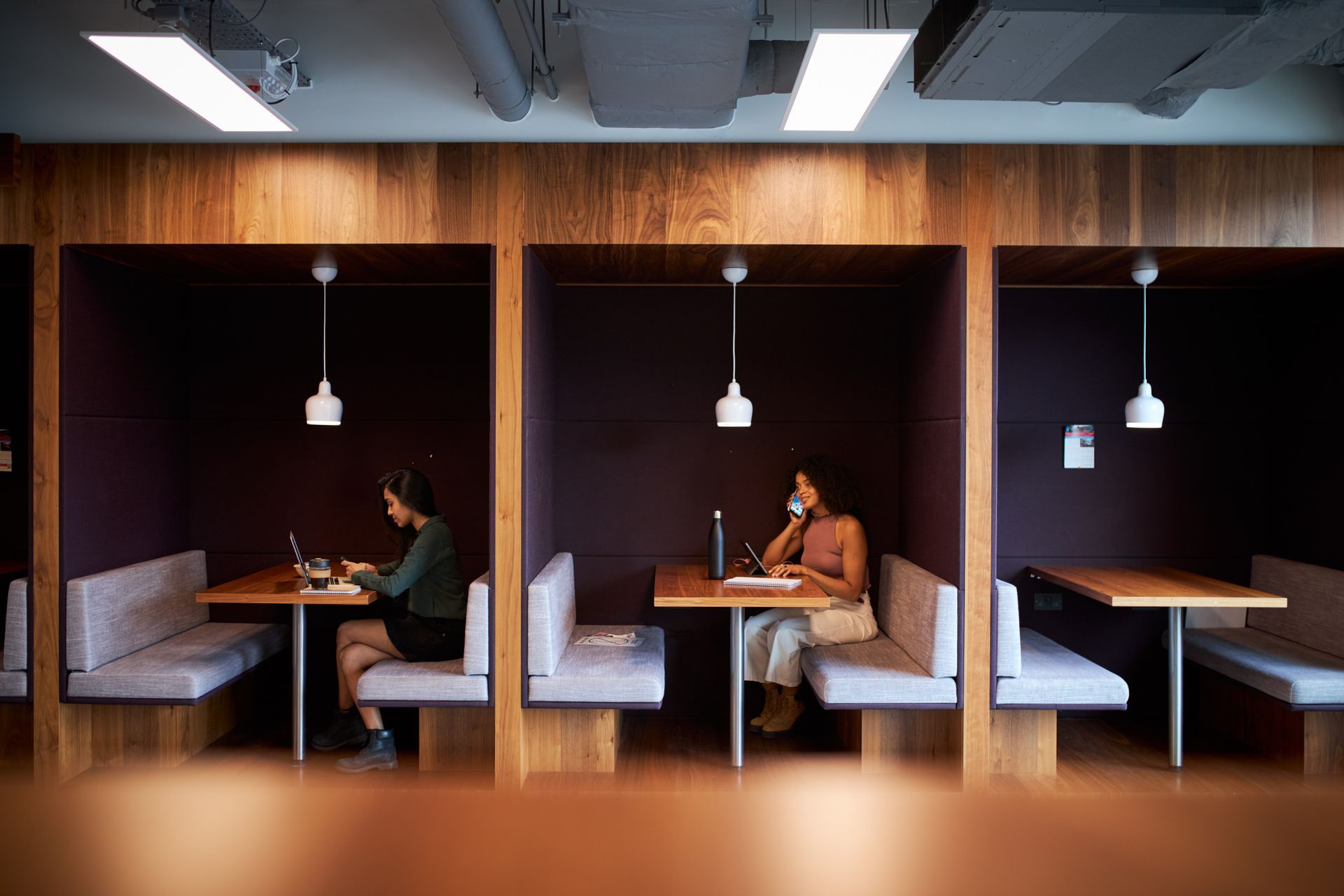 People working at desks in wood-paneled, enclosed booths. One uses a laptop, another talks on the phone.
