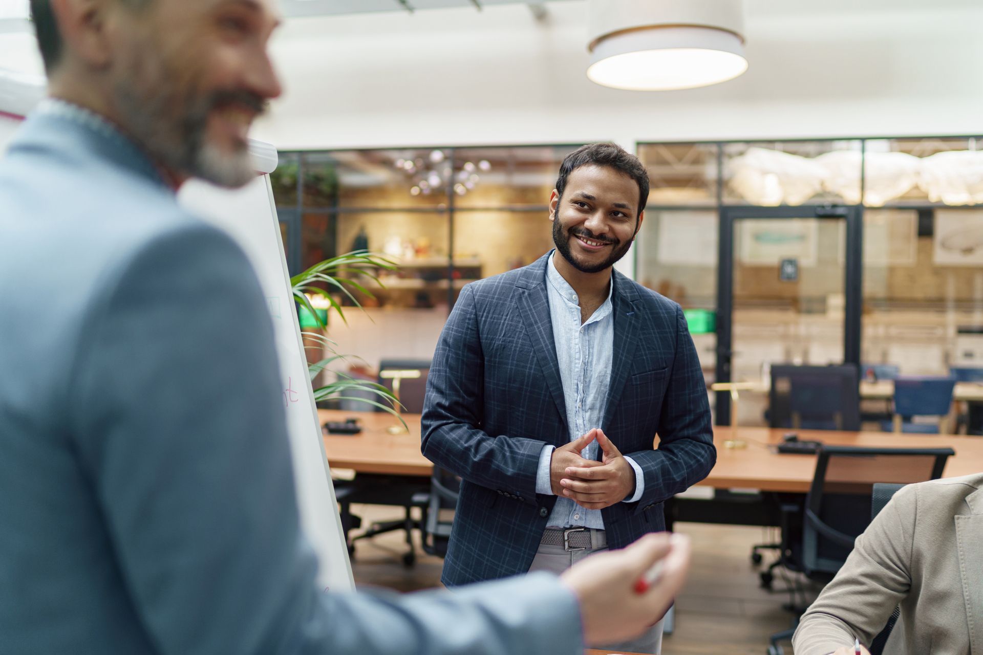 Two professionals in a modern office space, one smiling while gesturing during a presentation to a seated colleague.