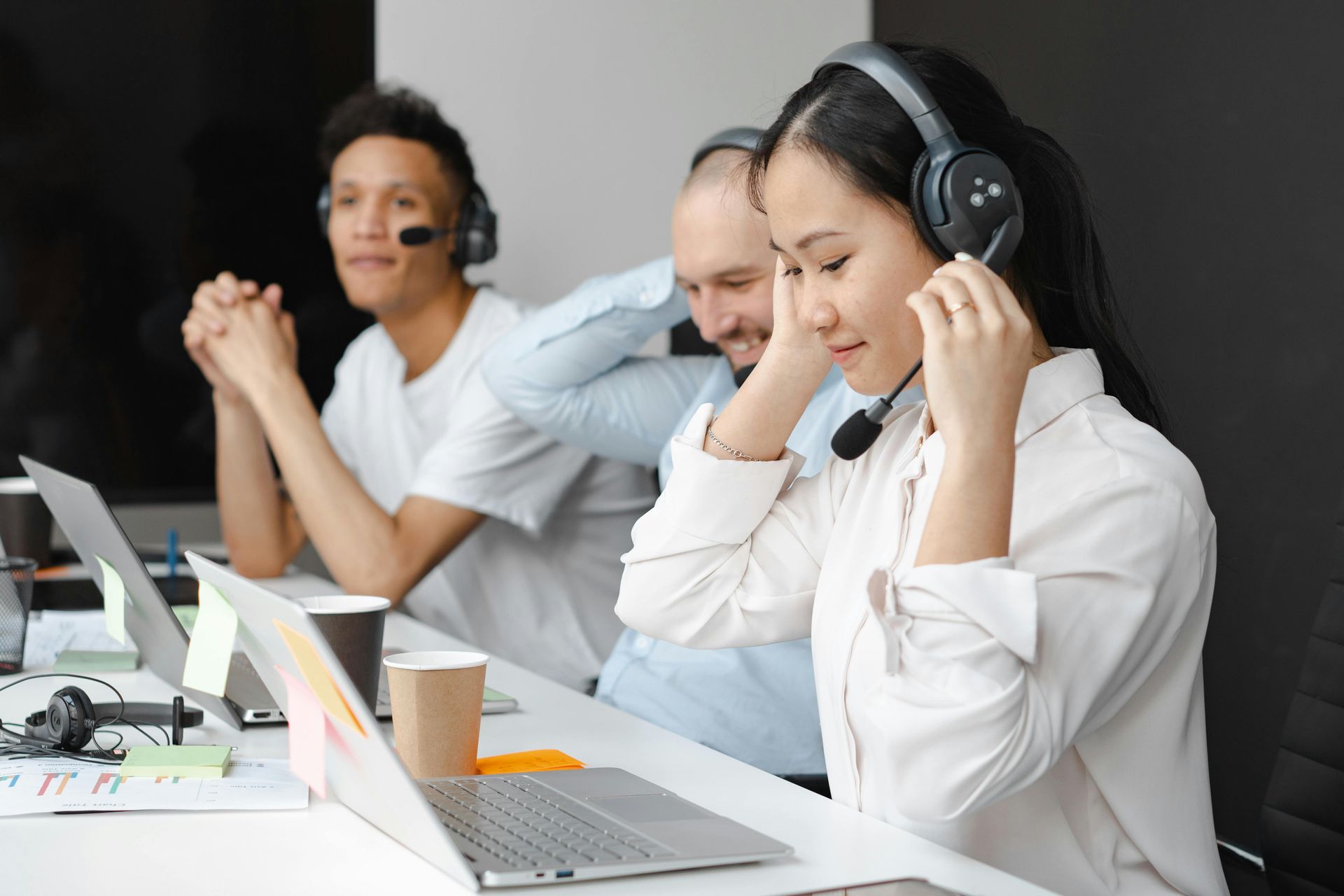 Three people wearing headsets at desks with laptops, working in an office environment.