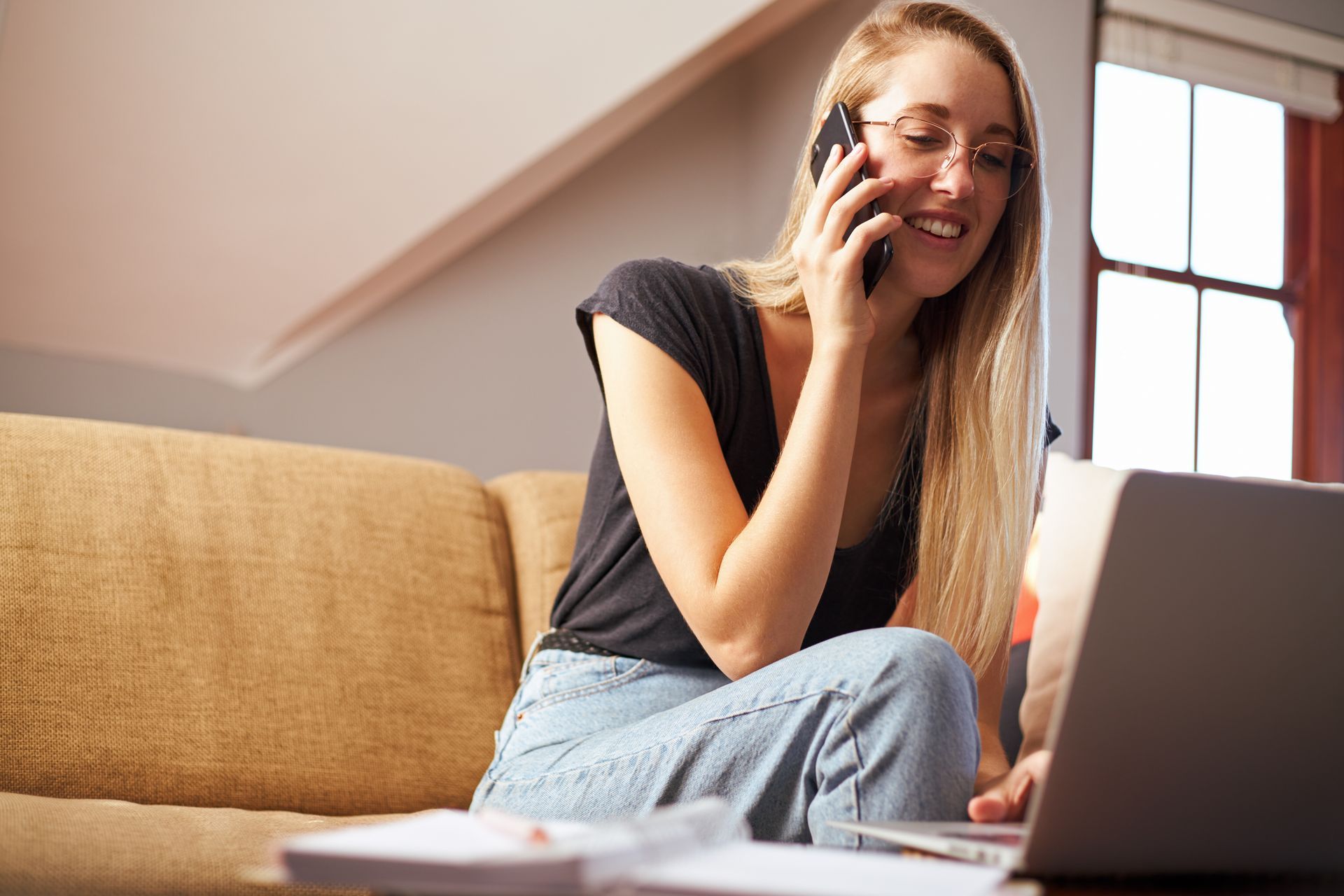 Woman on couch talking on phone, using laptop, smiling.