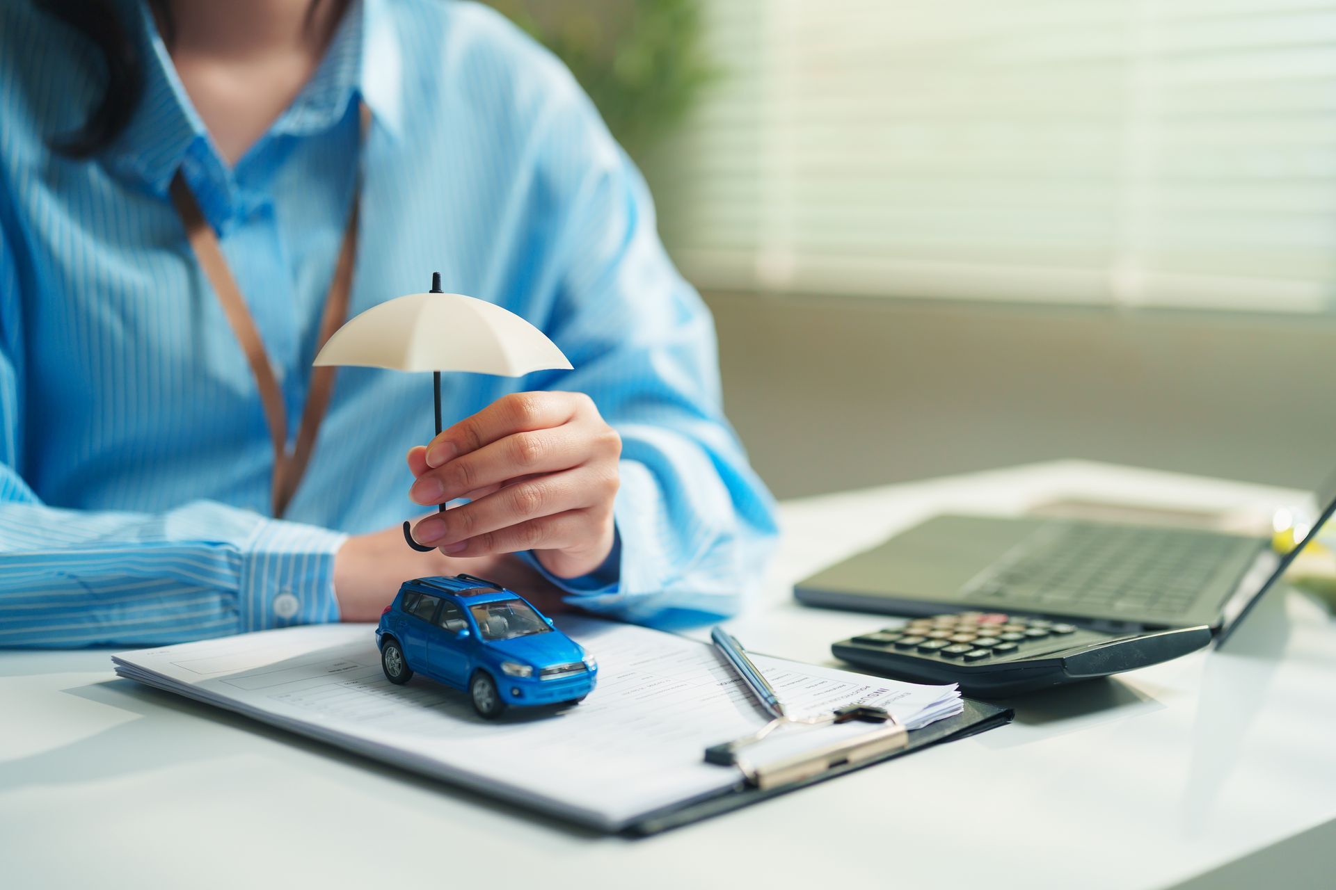 Person holding an umbrella over a toy car beside a laptop and calculator.