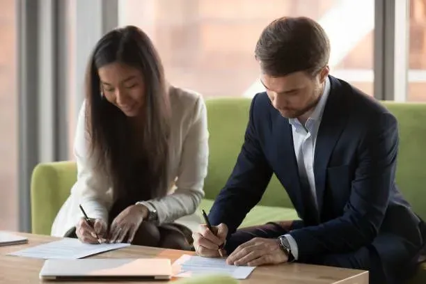 A woman and a man in business attire sign documents at a table, likely during a meeting. A woman and a man in business attire sign documents at a table, likely during a meeting.