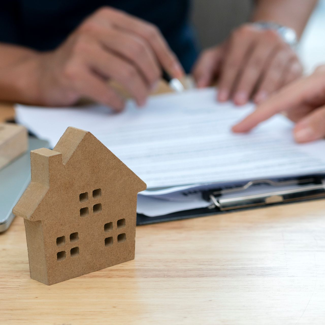 Wooden house model with hands pointing at and signing papers on a clipboard, likely a real estate transaction.