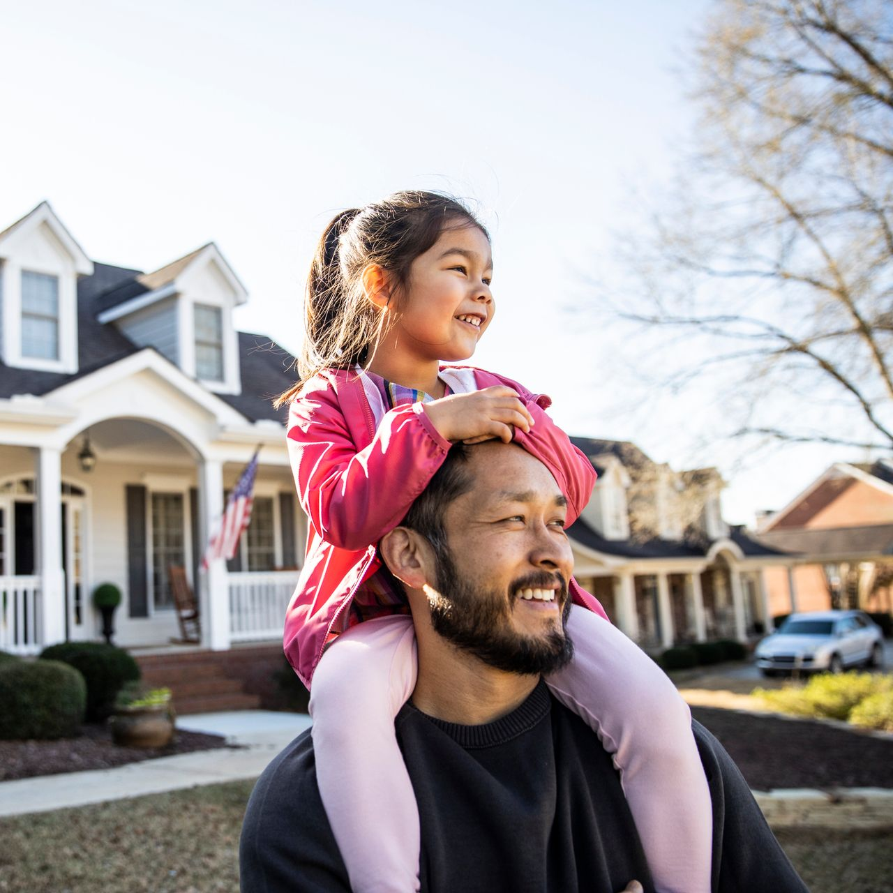 A man carries a young girl on his shoulders in front of a two-story house. The girl smiles and looks ahead.