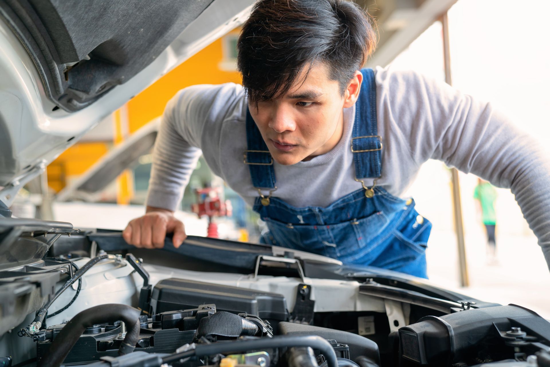 Mechanic in blue overalls examines car engine, focused expression. Outdoors, in a repair shop setting.