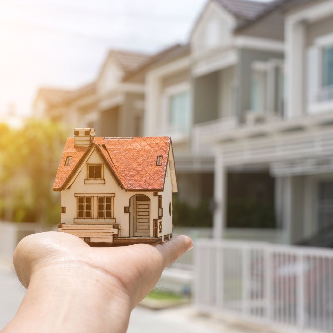 Hand holding a small wooden house in front of a row of suburban houses. Sunlight shines.