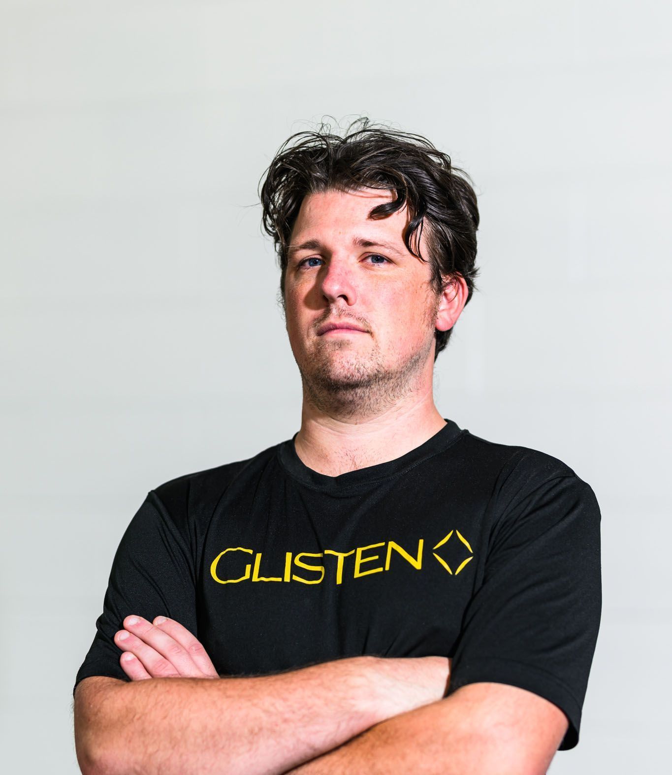 Man with crossed arms, wearing a black shirt with “GLISTEN” in gold lettering; neutral expression.