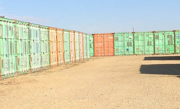Rows of green shipping containers in a dusty yard under a clear blue sky