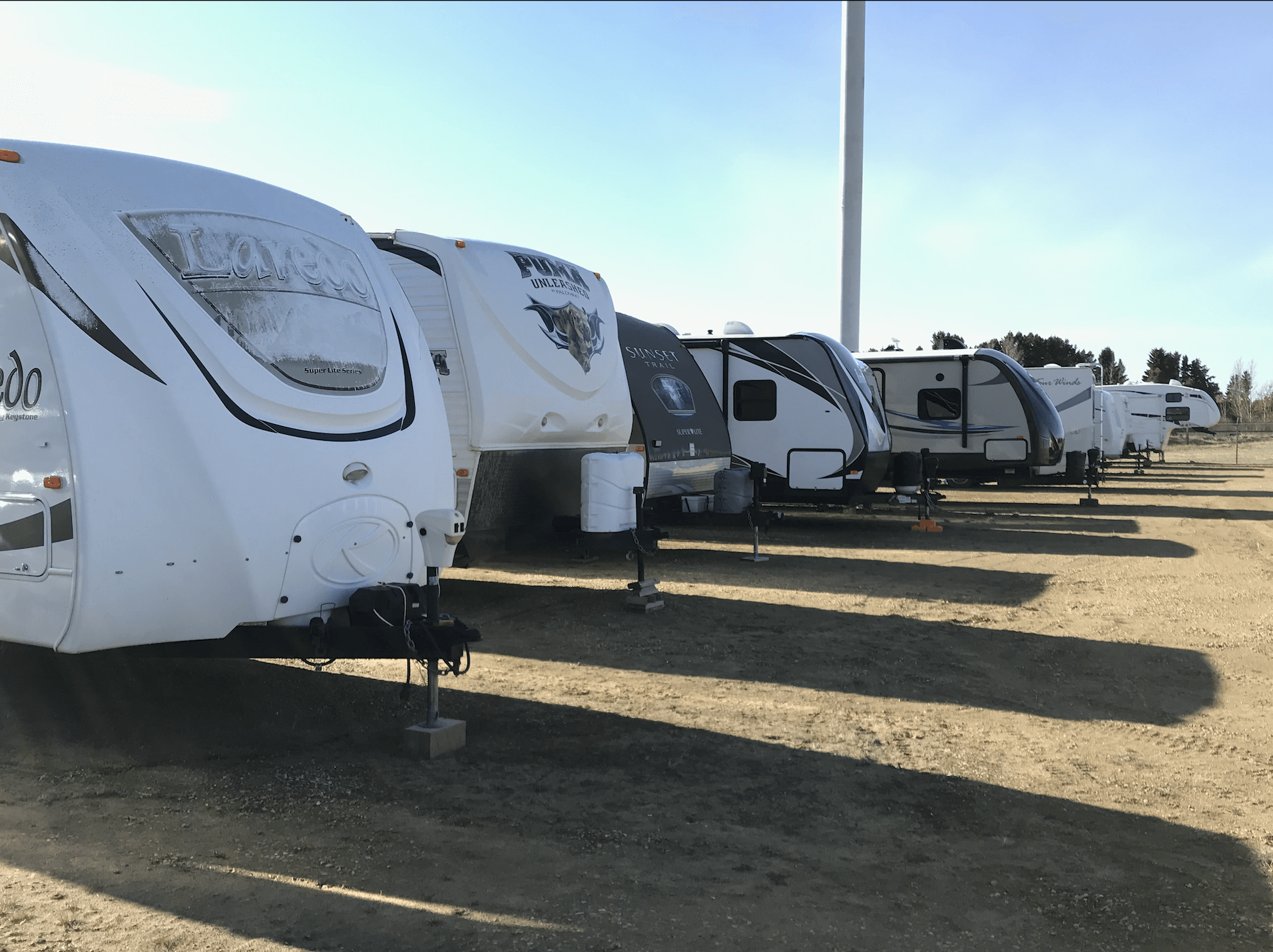 Row of white RV trailers parked on a dirt lot under a clear blue sky