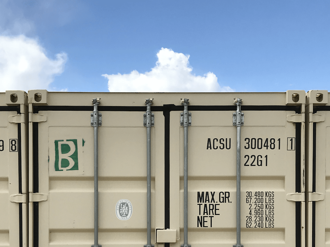 Beige shipping container against blue sky, with black markings and a green “B” logo.