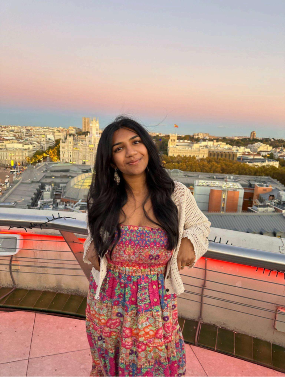 Woman in a floral dress standing on a rooftop at sunset, overlooking a city skyline.