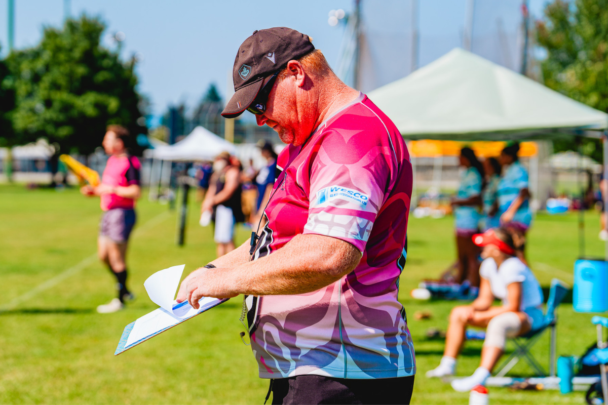 Man in a pink shirt and cap holding a clipboard at an outdoor sports event.