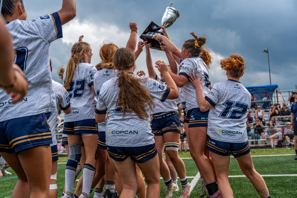 Women’s rugby team in white jerseys celebrates by lifting a trophy on the field